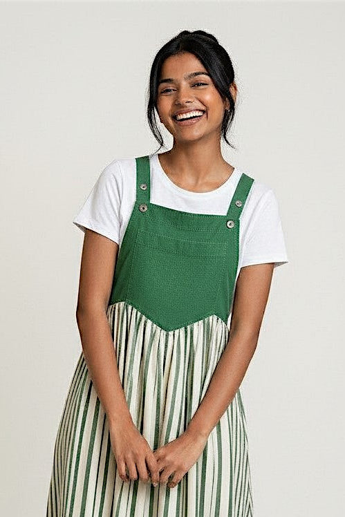 A woman modeling a green and white striped overall maxi dress over a white t-shirt, standing against a light gray studio background.
