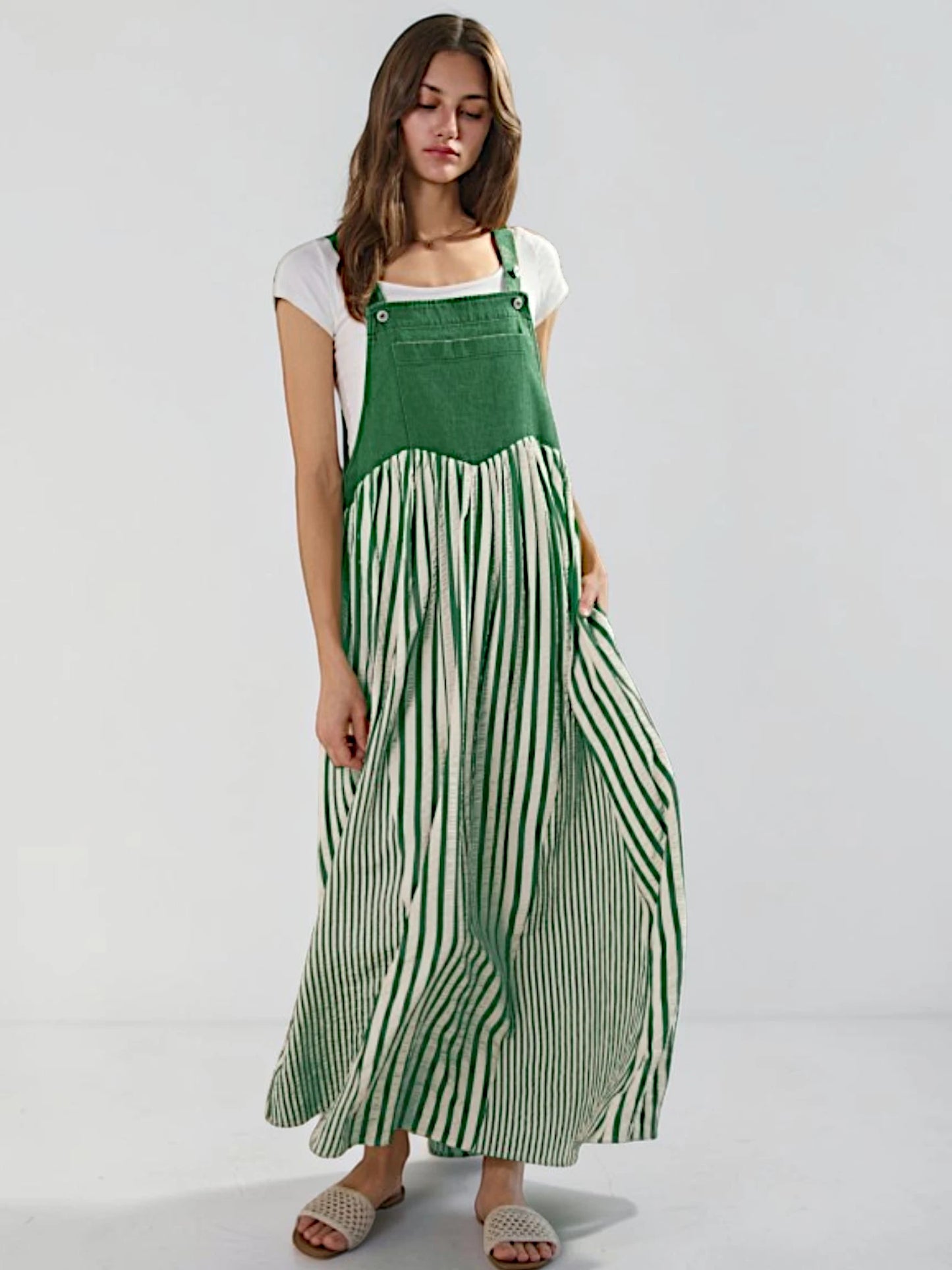 A woman modeling a green and white striped maxi pinafore dress over a white t-shirt, standing against a light gray studio background.