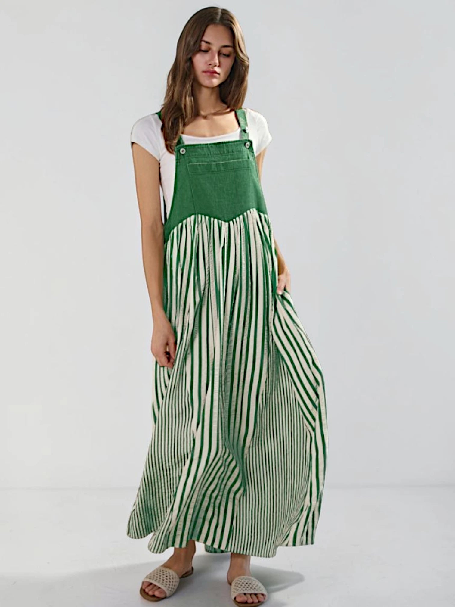 A woman modeling a green and white striped maxi pinafore dress over a white t-shirt, standing against a light gray studio background.