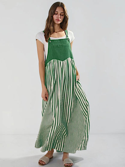 A woman modeling a green and white striped maxi pinafore dress over a white t-shirt, standing against a light gray studio background.