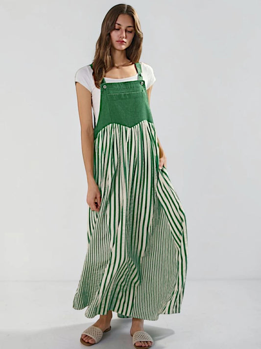 A woman modeling a green and white striped maxi pinafore dress over a white t-shirt, standing against a light gray studio background.