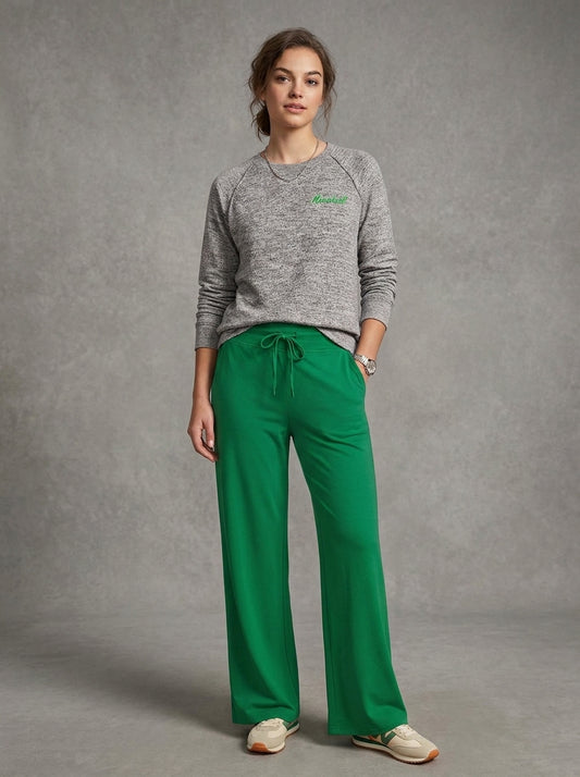 A woman modeling a heather gray long-sleeve crewneck sweatshirt with a small green embroidered logo on the chest, paired with bright green wide-leg drawstring pants and beige sneakers, set against a textured gray studio backdrop.