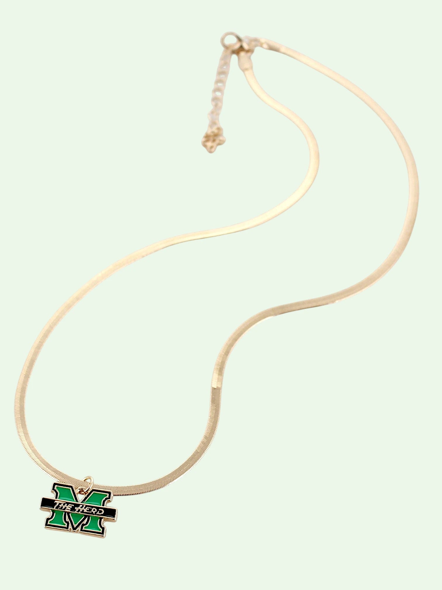 A gold-toned snake chain necklace featuring a green and black enamel 'M' pendant with 'The HERD' text, displayed against a pale mint green background.