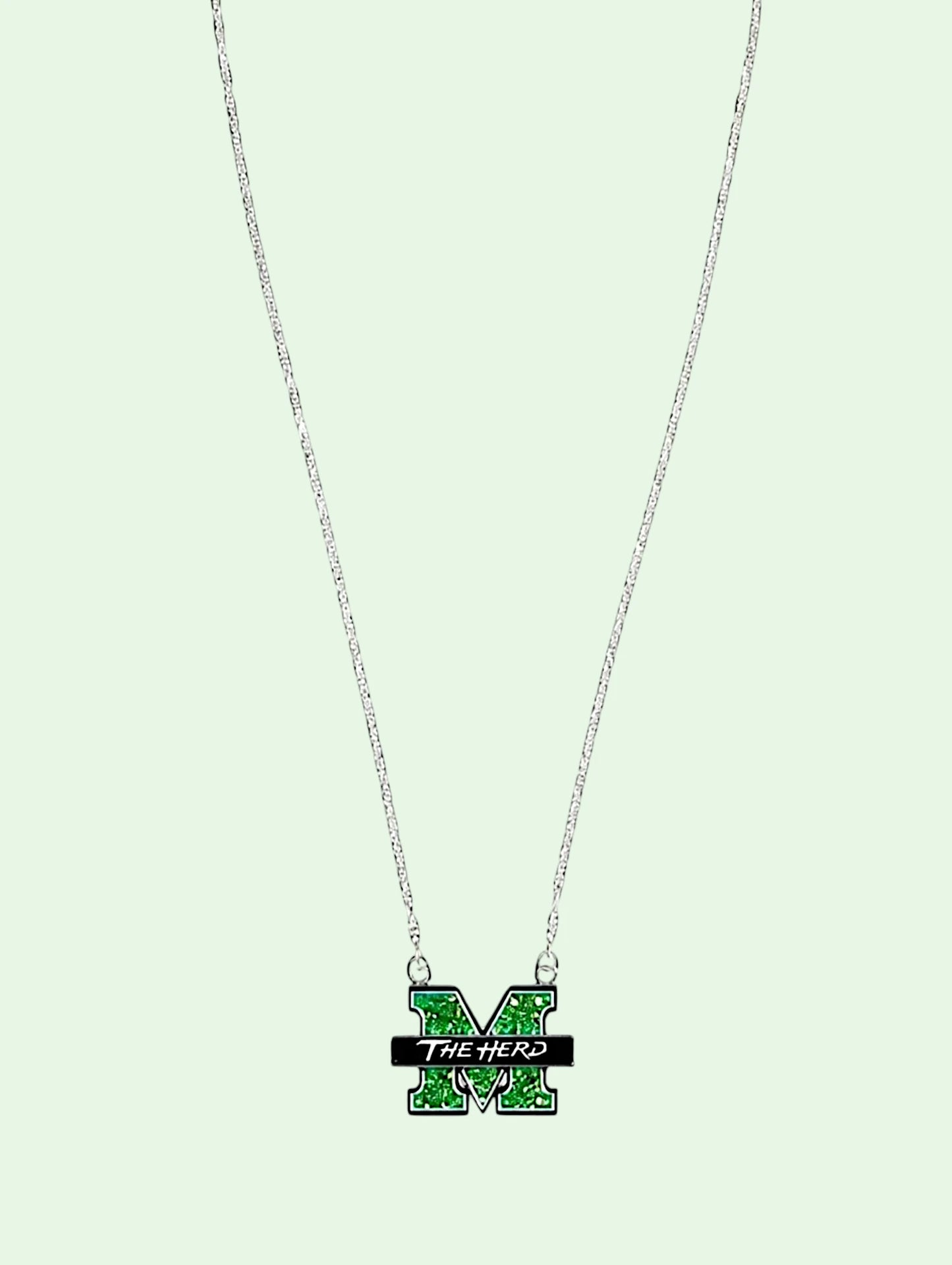 A silver chain necklace featuring a green and black enamel 'M' pendant with the text 'THE HERD' on a light mint green background.