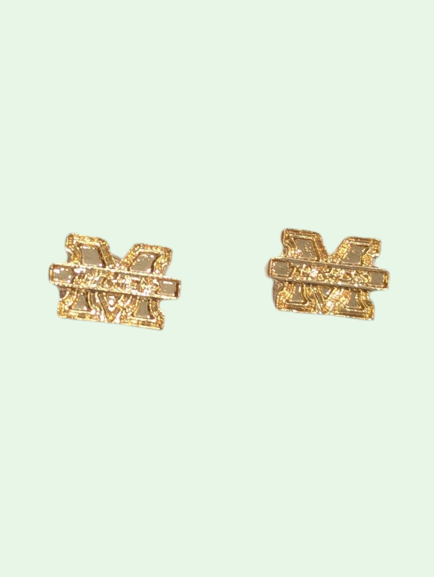 A pair of gold-toned stud earrings shaped like the letter 'M' with a textured, glittery finish, set against a solid pale mint green background.