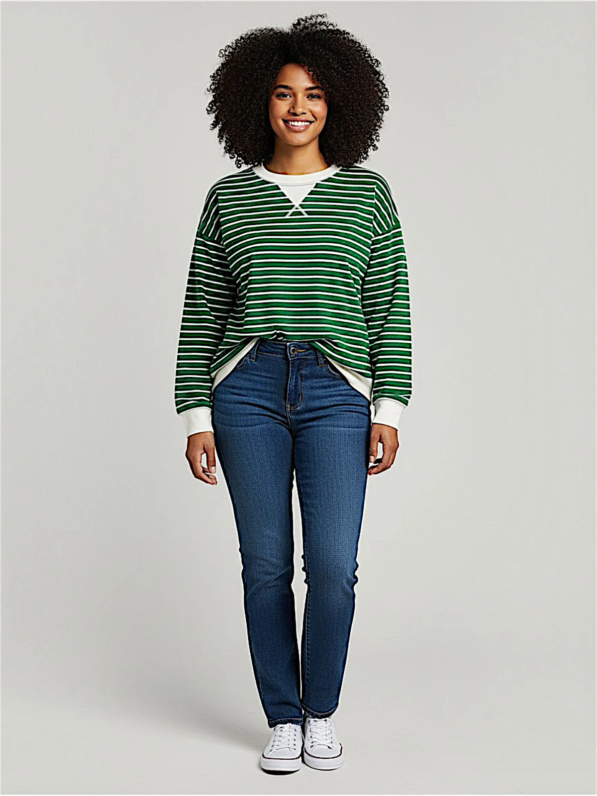 A woman with an afro hairstyle stands against a light gray studio background wearing a green and white striped long-sleeve sweater, blue jeans, and white sneakers.