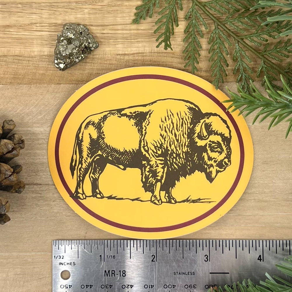 Round yellow sticker with a bison design on a wooden surface with a ruler for scale.