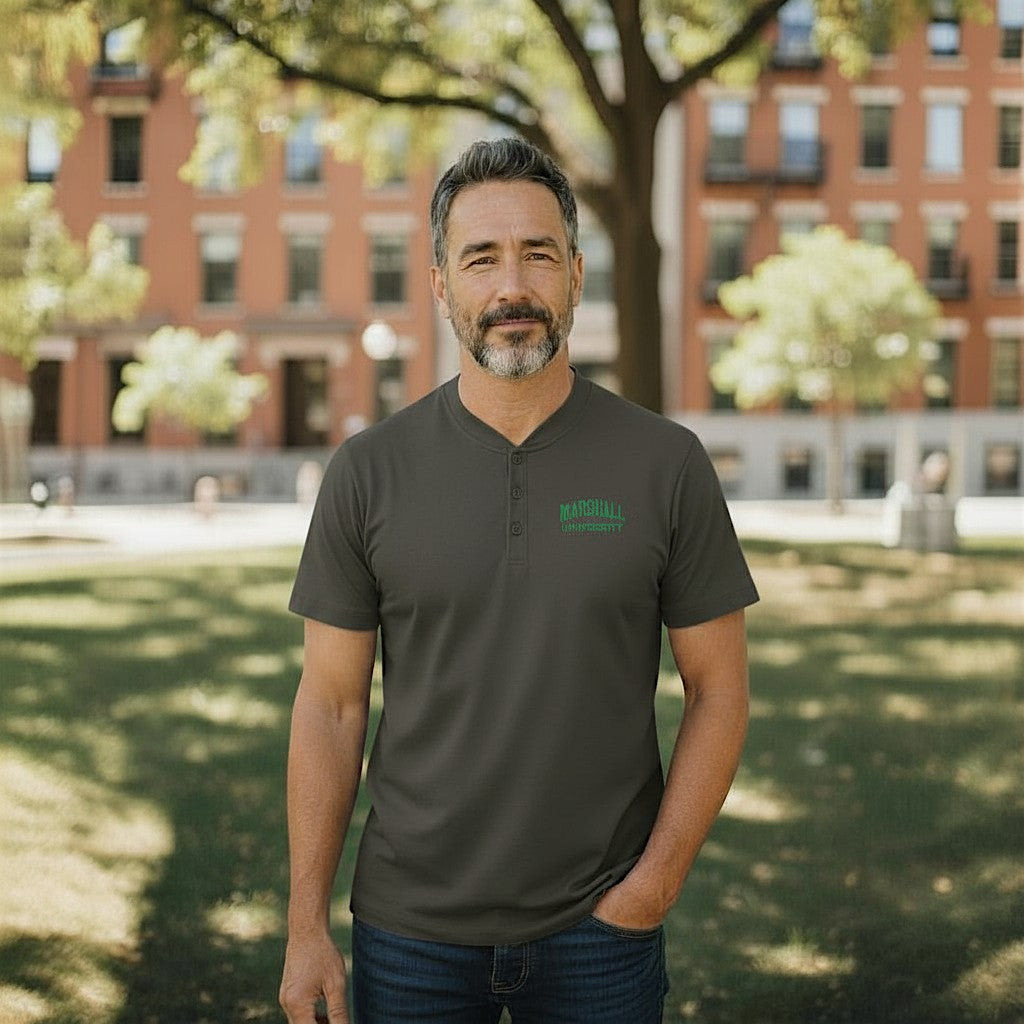 Man wearing a grey polo shirt with a logo, standing in a park with trees and buildings in the background.