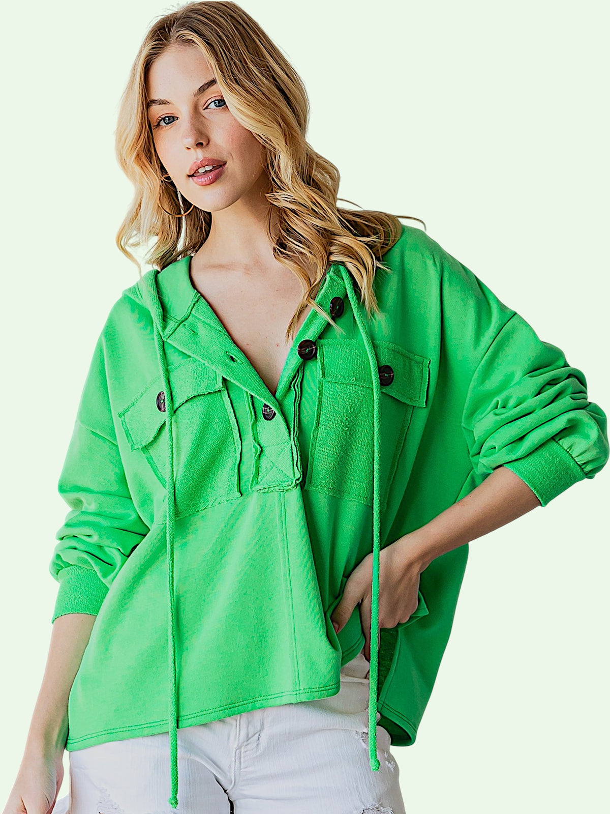 A woman wearing a bright green cropped hooded button-up shirt with drawstrings and front pockets, paired with white pants, against a pale green studio background.