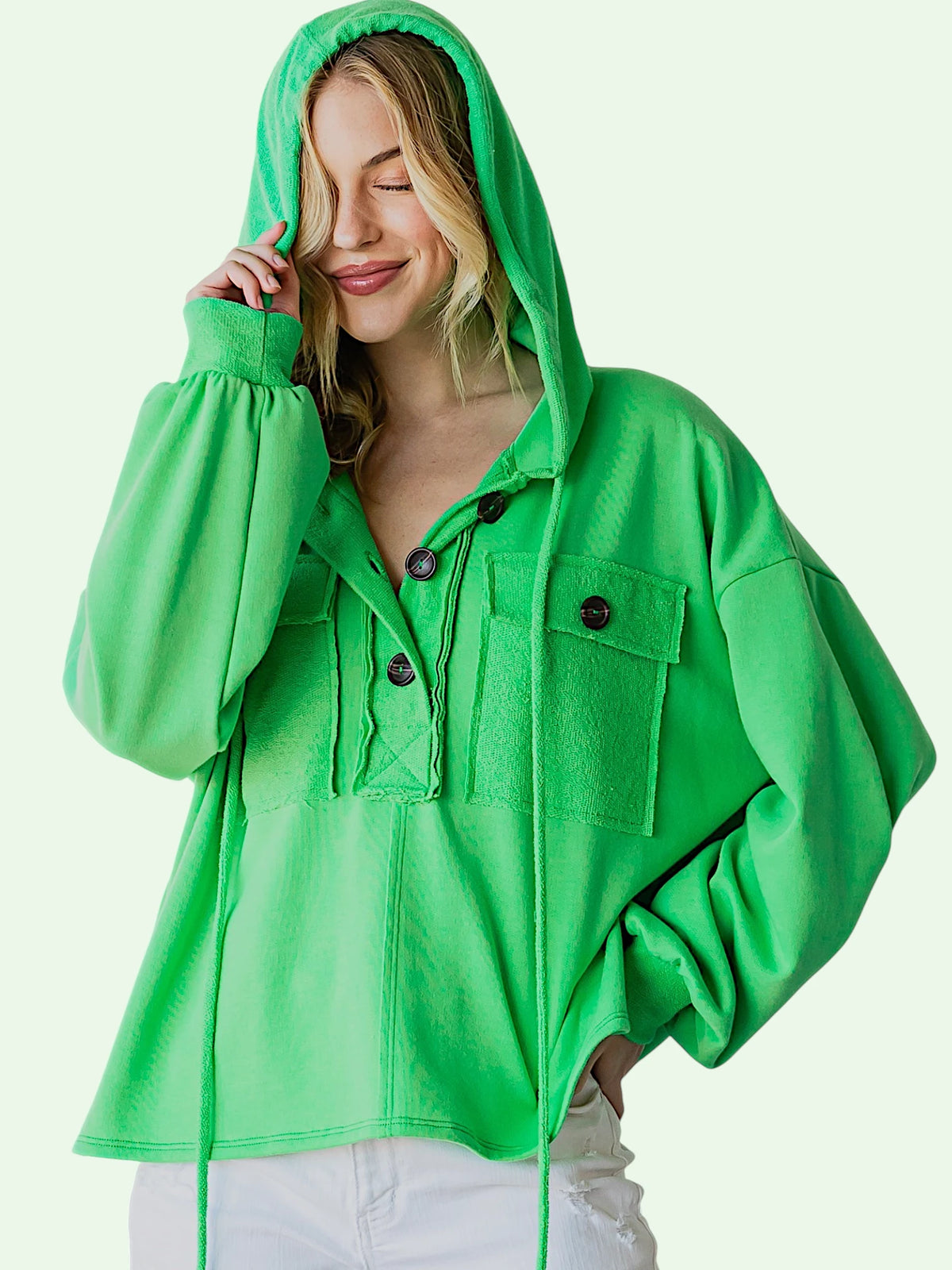 A woman wearing a bright green hooded pullover jacket with front flap pockets and drawstrings, paired with white pants, against a pale mint background.