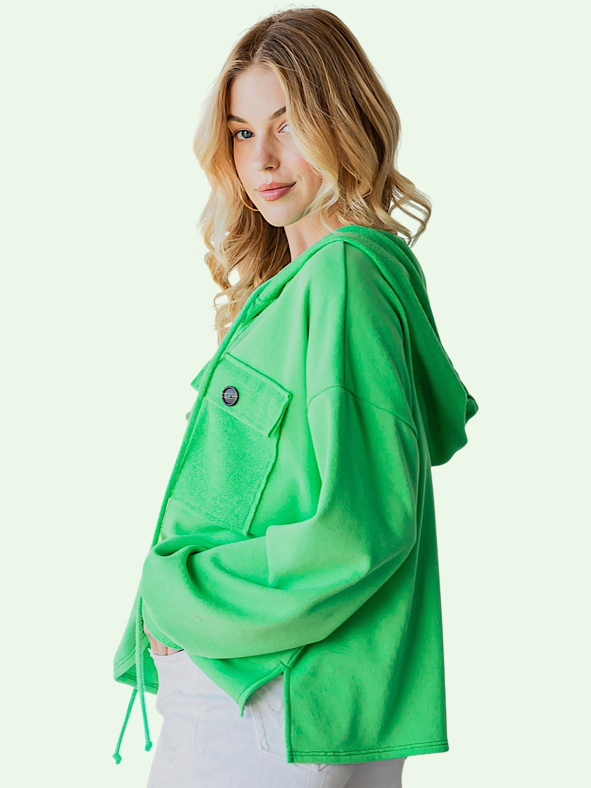 A woman wearing a bright green cropped hooded jacket with a buttoned chest pocket, shown against a pale mint background.