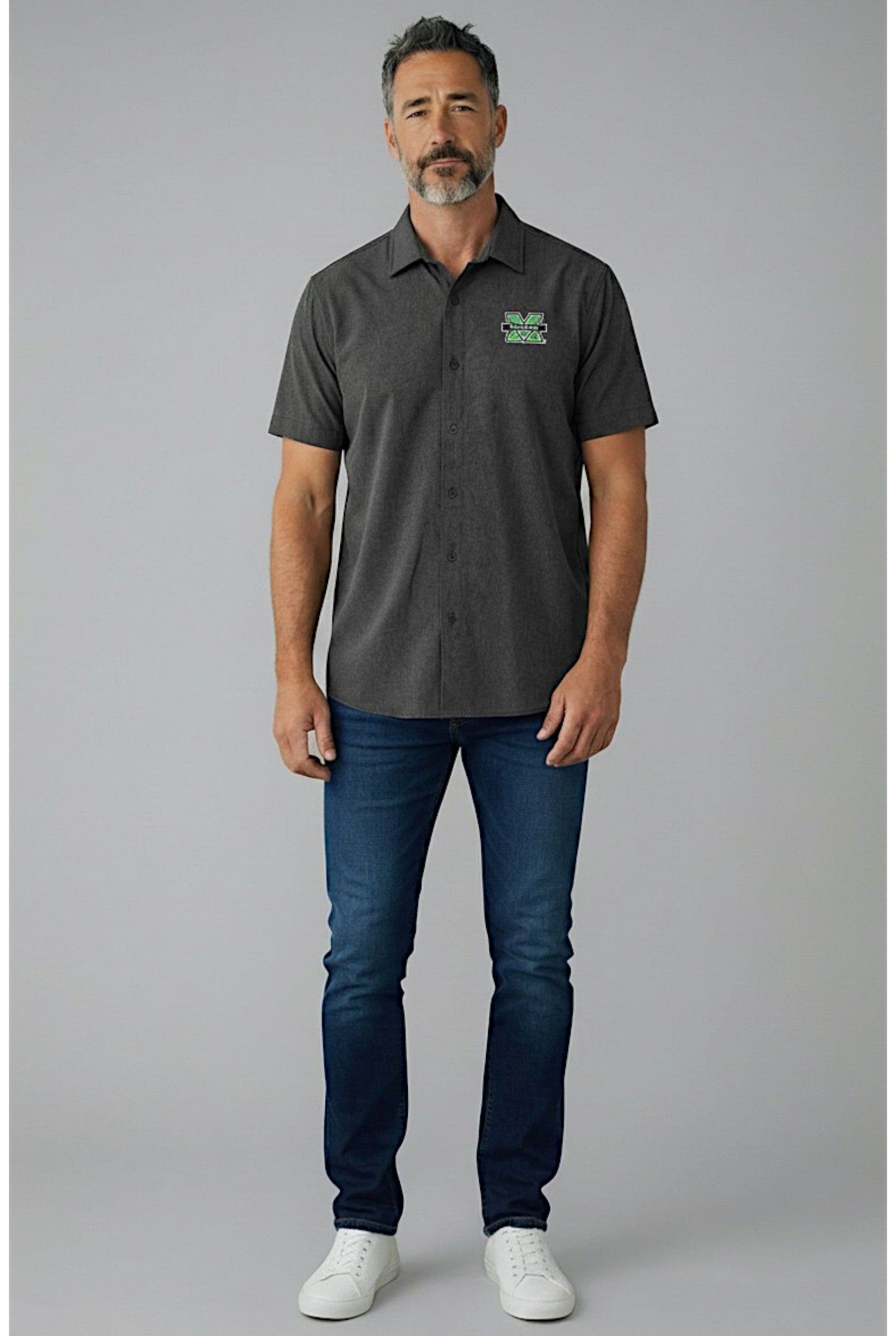 Man wearing a dark grey camp shirt with a logo and blue jeans on a gray background