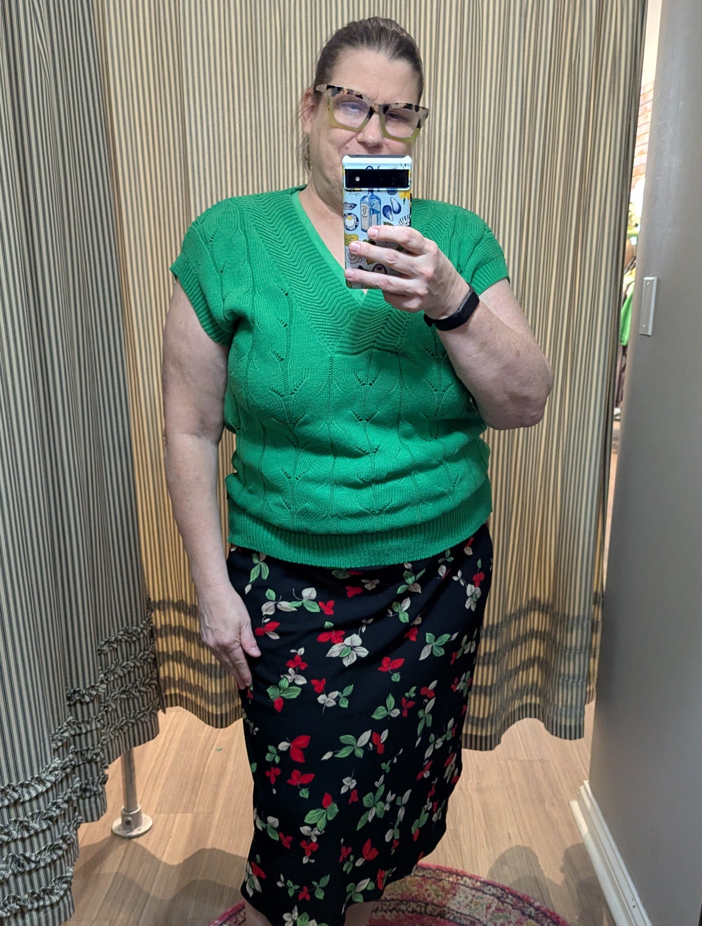 A woman takes a mirror selfie in a fitting room wearing a bright green short-sleeve knit top and a black floral skirt with red and green accents.