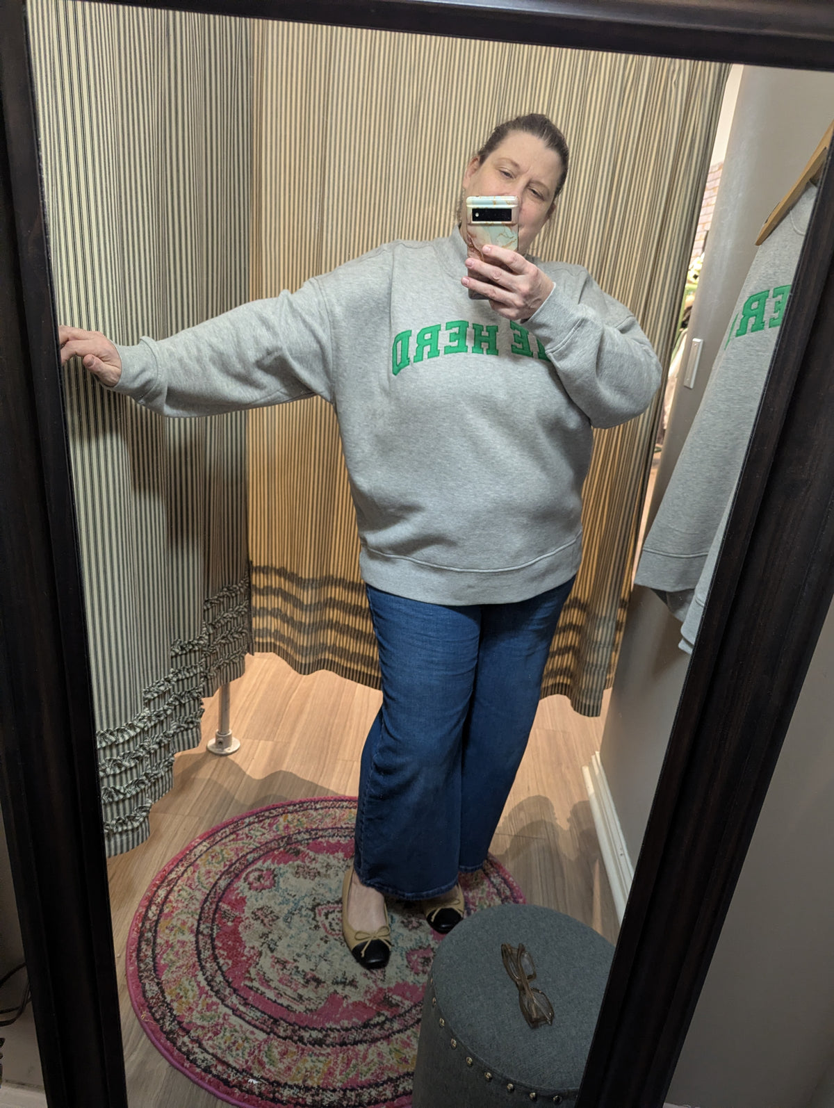 A woman takes a mirror selfie in a fitting room wearing a light gray oversized sweatshirt with green text reading 'THE HERD' and blue jeans.