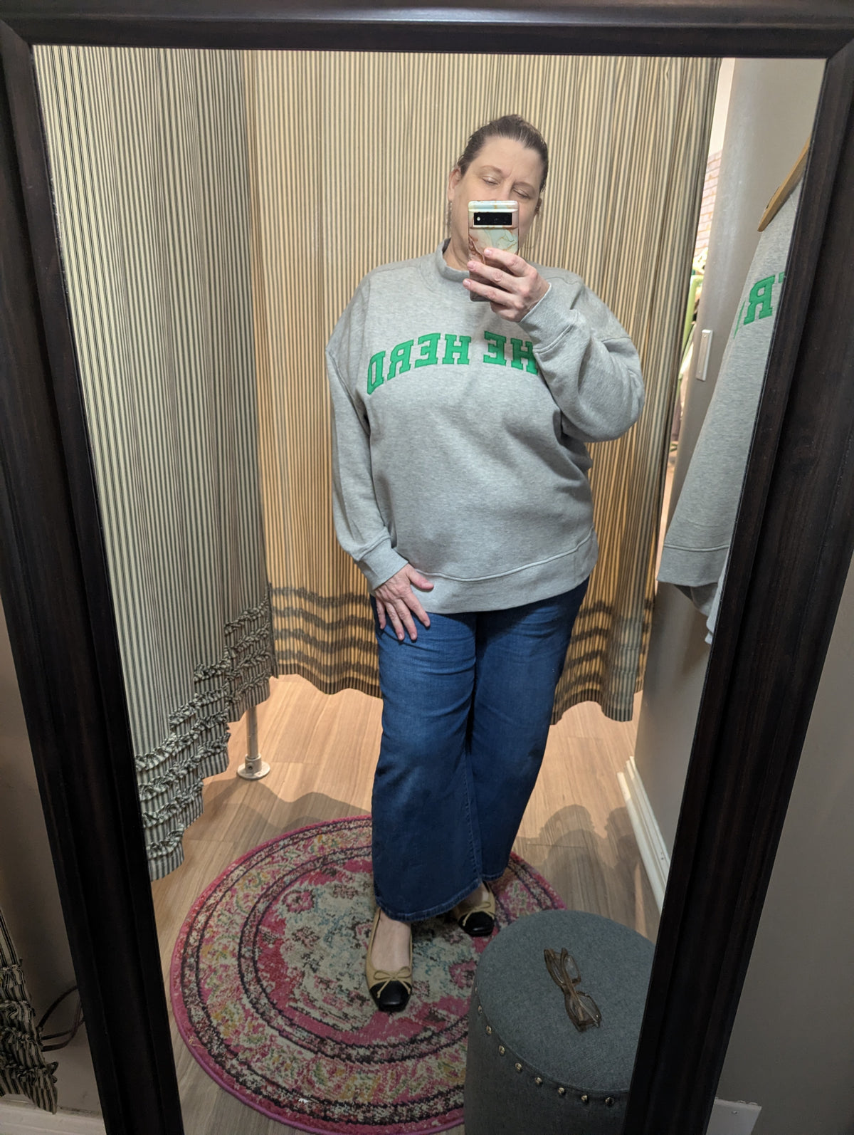 A person taking a mirror selfie in a fitting room wearing a heather gray sweatshirt with green text reading 'THE HEAD' backwards, blue wide-leg jeans, and black flat shoes.