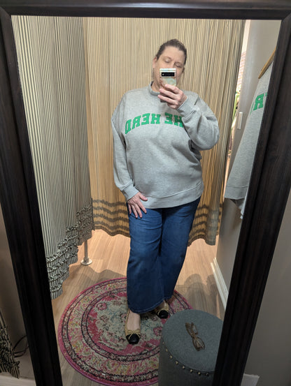 A person taking a mirror selfie in a fitting room wearing a heather gray sweatshirt with green text reading 'THE HEAD' backwards, blue wide-leg jeans, and black flat shoes.