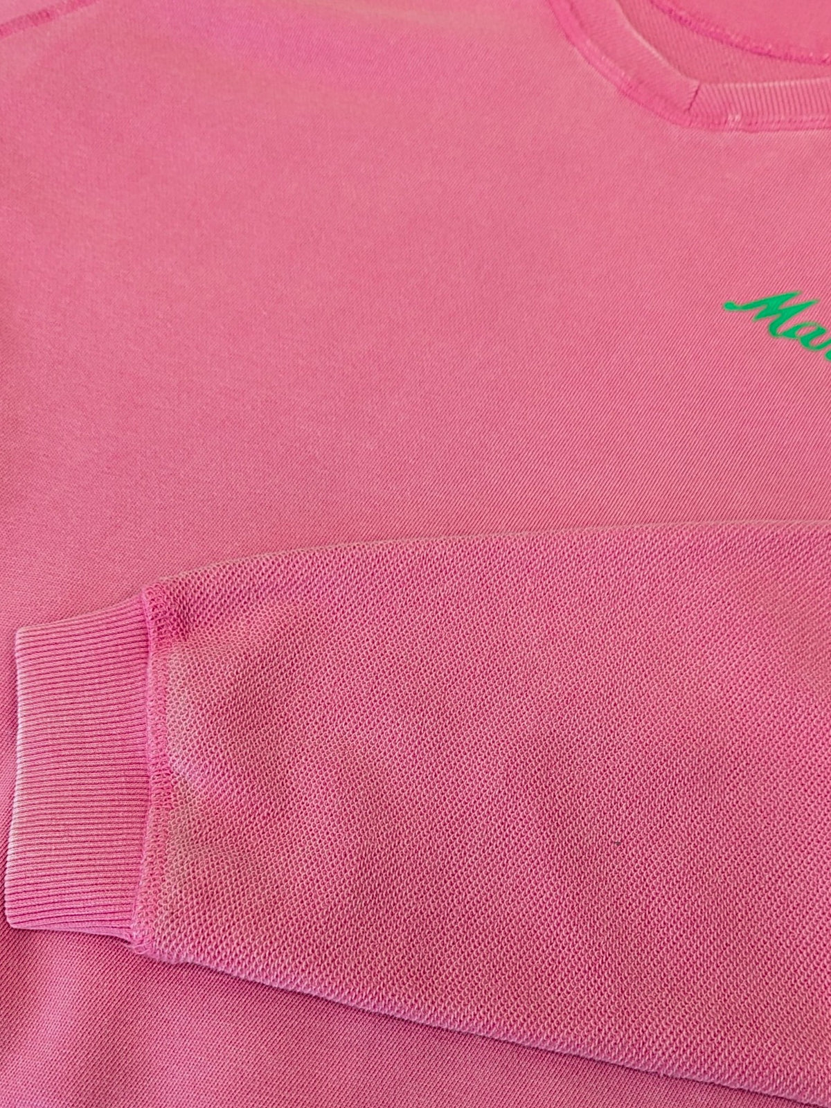 Close-up of a pink crewneck sweatshirt showing the ribbed cuff of the left sleeve and a small green embroidered script logo on the chest.