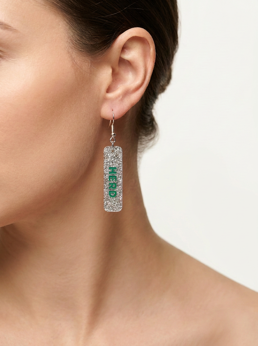 A close-up of a model's ear wearing a silver rectangular drop earring covered in clear rhinestones with the word 'NERD' written vertically in green.