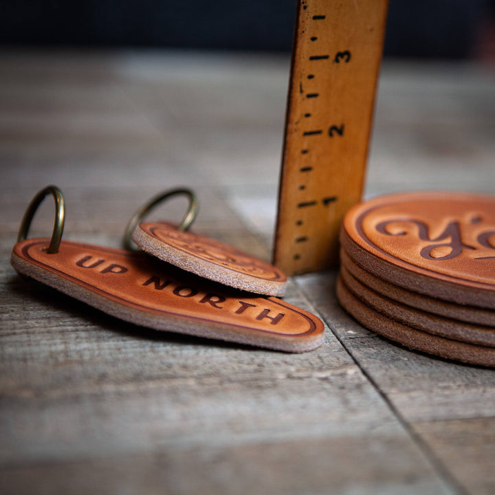 Leather keychains and coasters with text on a wooden surface, accompanied by a ruler for scale.