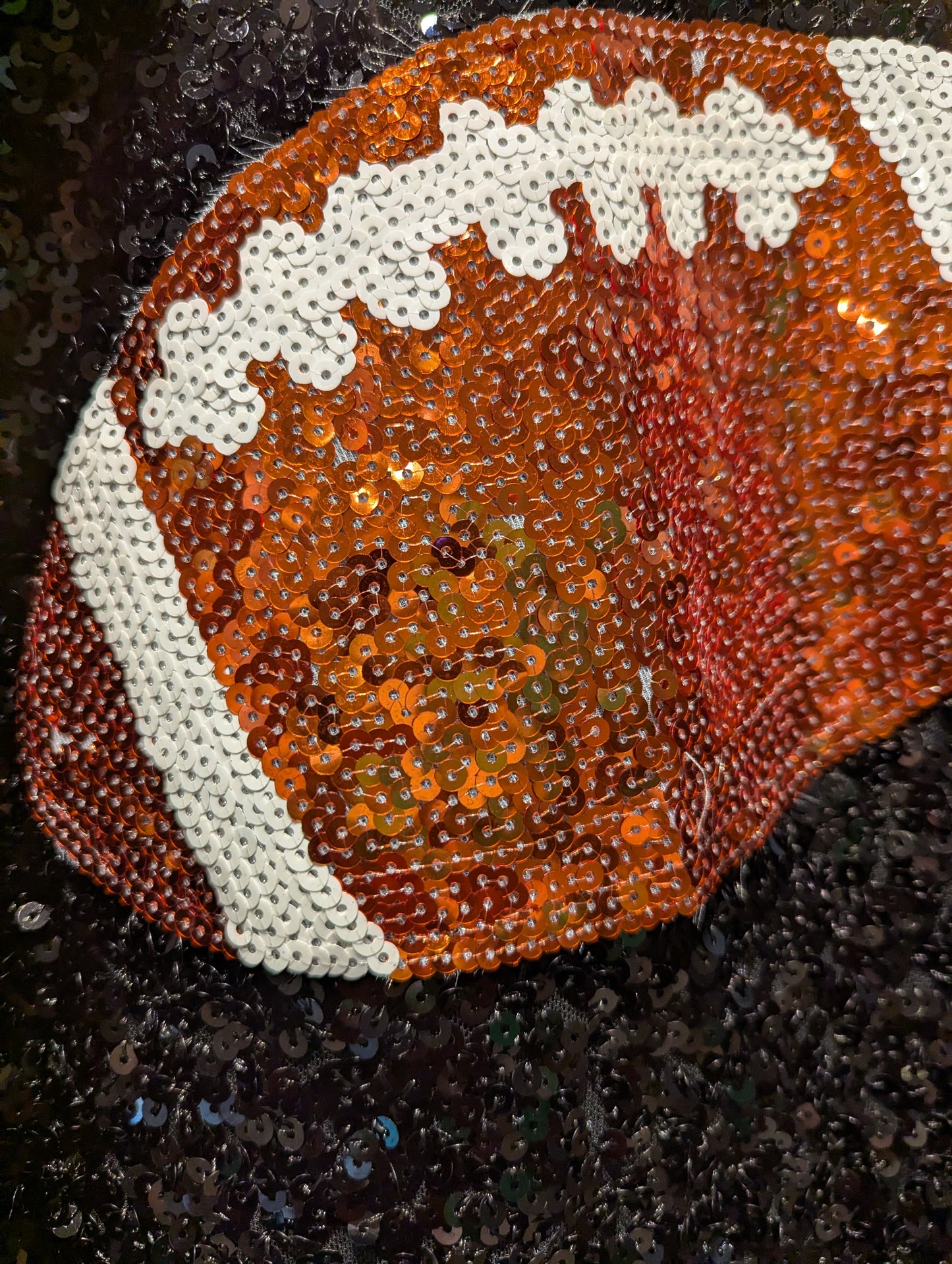 A close-up of a football-shaped sequin accessory, likely a hat or pouch, featuring an orange and brown pattern with white laces and stripes, set against a dark sequined background.