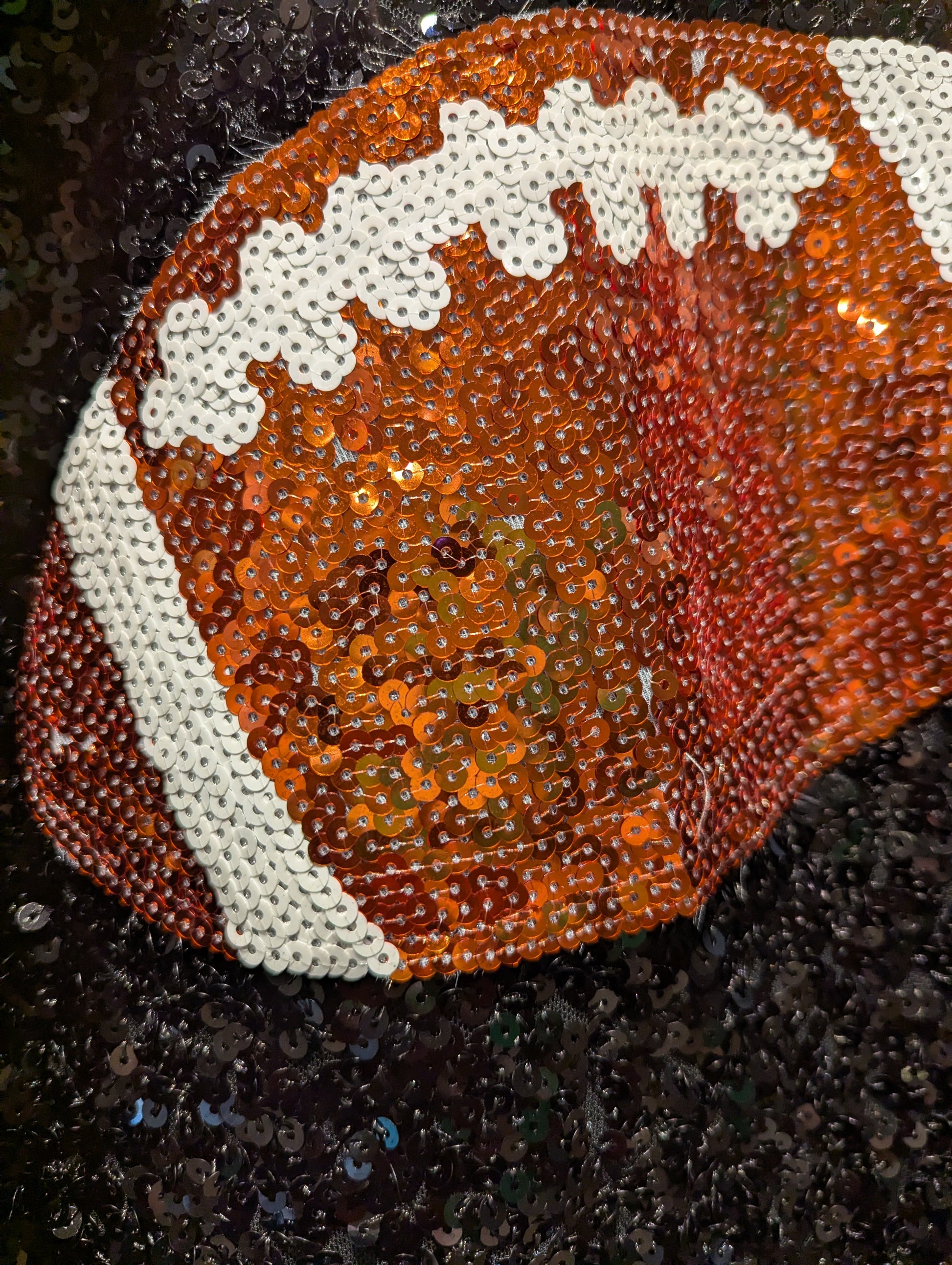 A close-up of a football-shaped sequin accessory, likely a hat or pouch, featuring an orange and brown pattern with white laces and stripes, set against a dark sequined background.