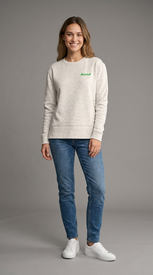 A woman modeling a light heather gray crewneck sweatshirt with a green printed 
'Marshall' logo on the chest, paired with blue jeans and white sneakers, against a solid gray studio background.