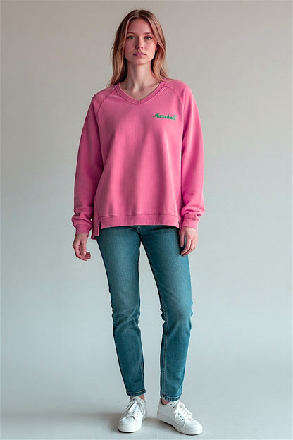 A woman modeling a pink v-neck sweatshirt with a small green printed Marshall logo on the left chest, paired with blue jeans and white sneakers, against a plain light gray studio background.