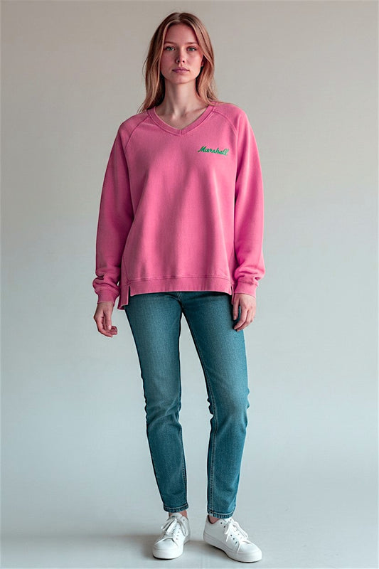 A woman modeling a pink v-neck sweatshirt with a small green printed Marshall logo on the left chest, paired with blue jeans and white sneakers, against a plain light gray studio background.