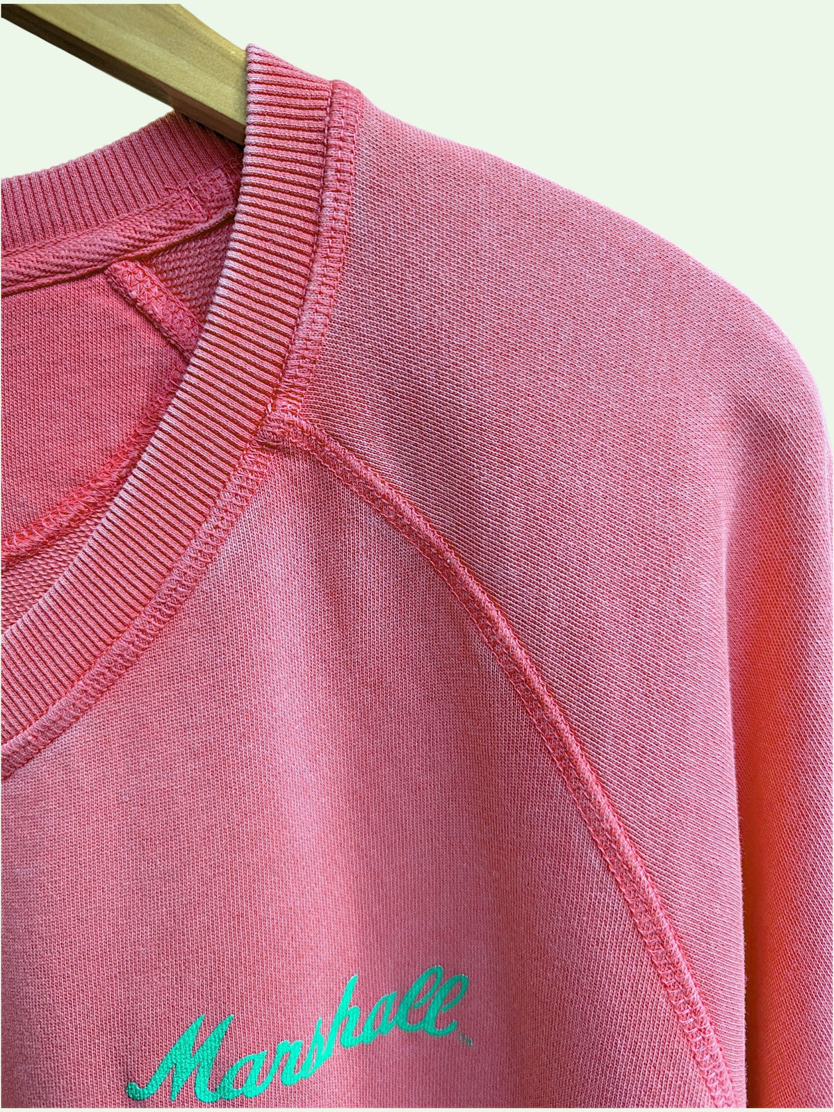 A close-up of a pink crewneck sweatshirt hanging on a wooden hanger, featuring a bright green embroidered 'Marshall' logo on the chest.