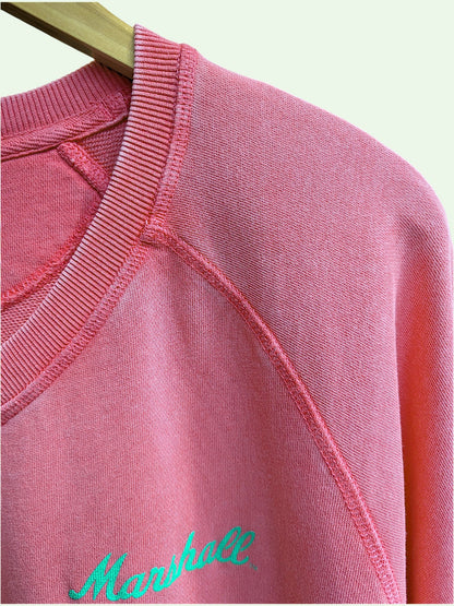 A close-up of a pink crewneck sweatshirt hanging on a wooden hanger, featuring a bright green embroidered 'Marshall' logo on the chest.