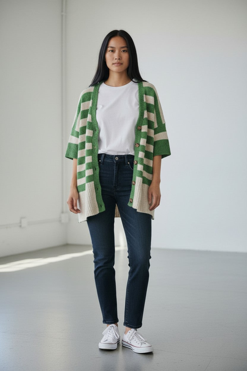 Person wearing a green and white striped cardigan, white shirt, dark jeans, and white sneakers in a minimalistic setting.
