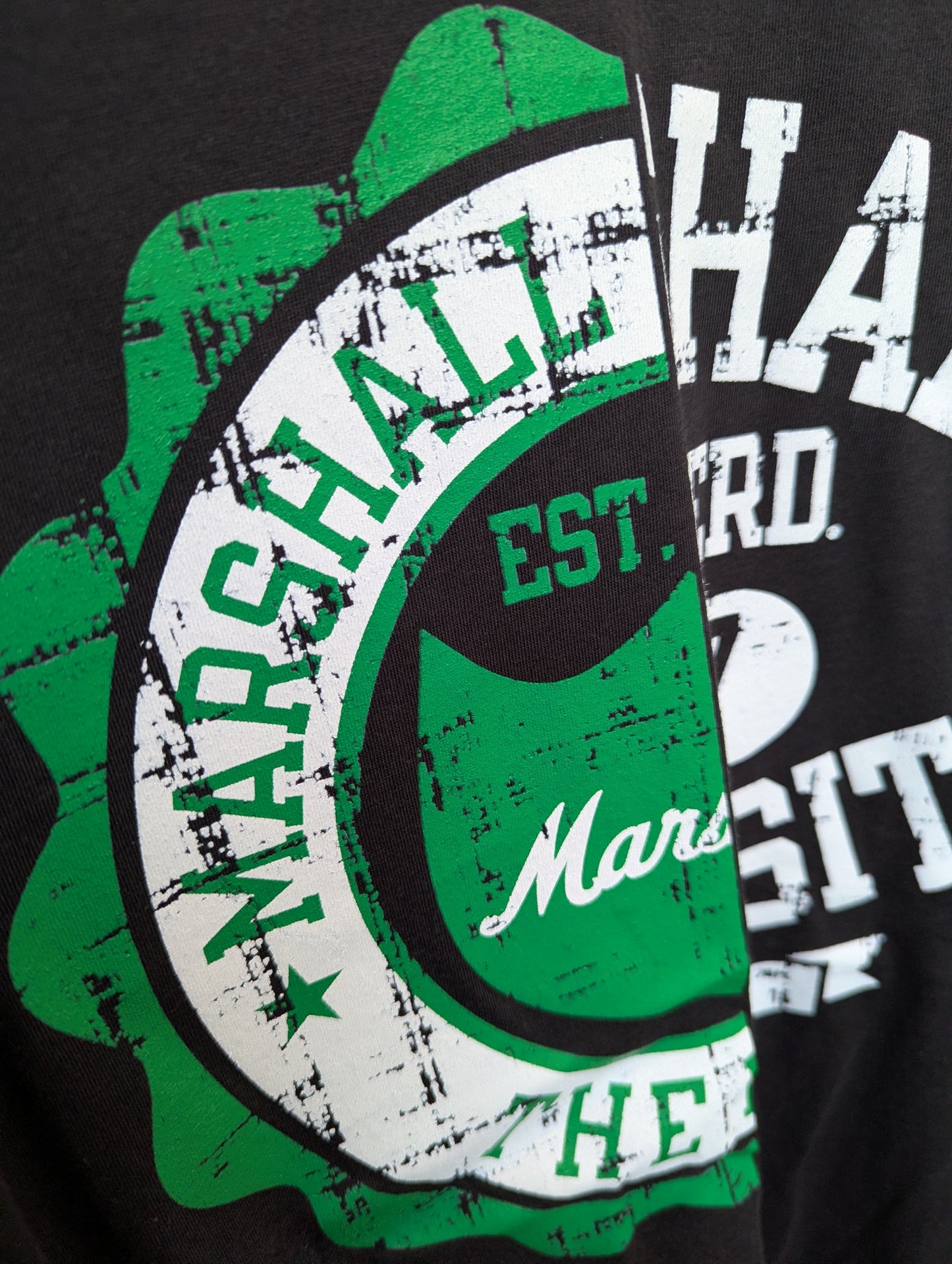 Close-up of a black graphic t-shirt featuring a distressed green and white circular badge design with the text 'MARSHALL HEAD' and 'EST. 2025'.