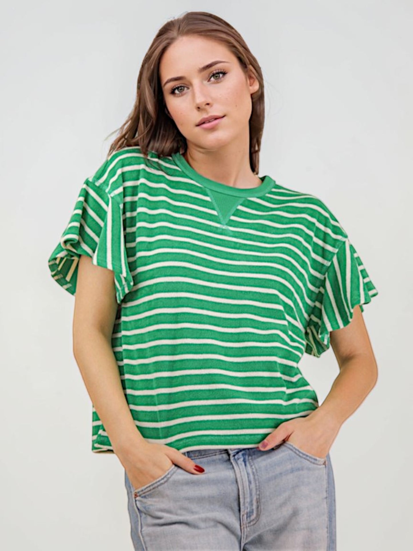 A woman wearing a green and white horizontally striped short-sleeve top with ruffled sleeves, paired with light-wash jeans, against a plain light gray background.