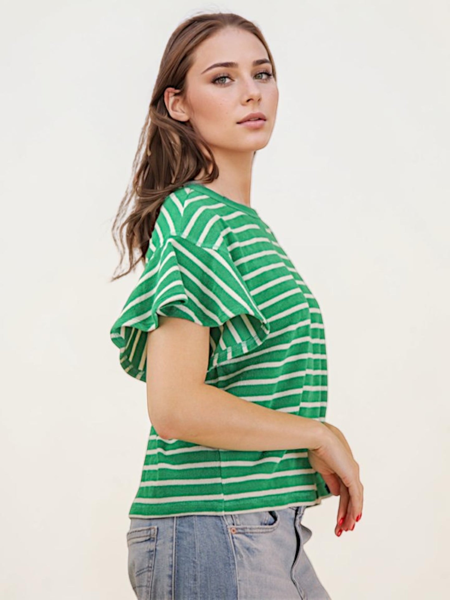 A woman wearing a green and white striped short-sleeve top with ruffled sleeves, paired with light blue jeans, photographed against a plain off-white studio background.