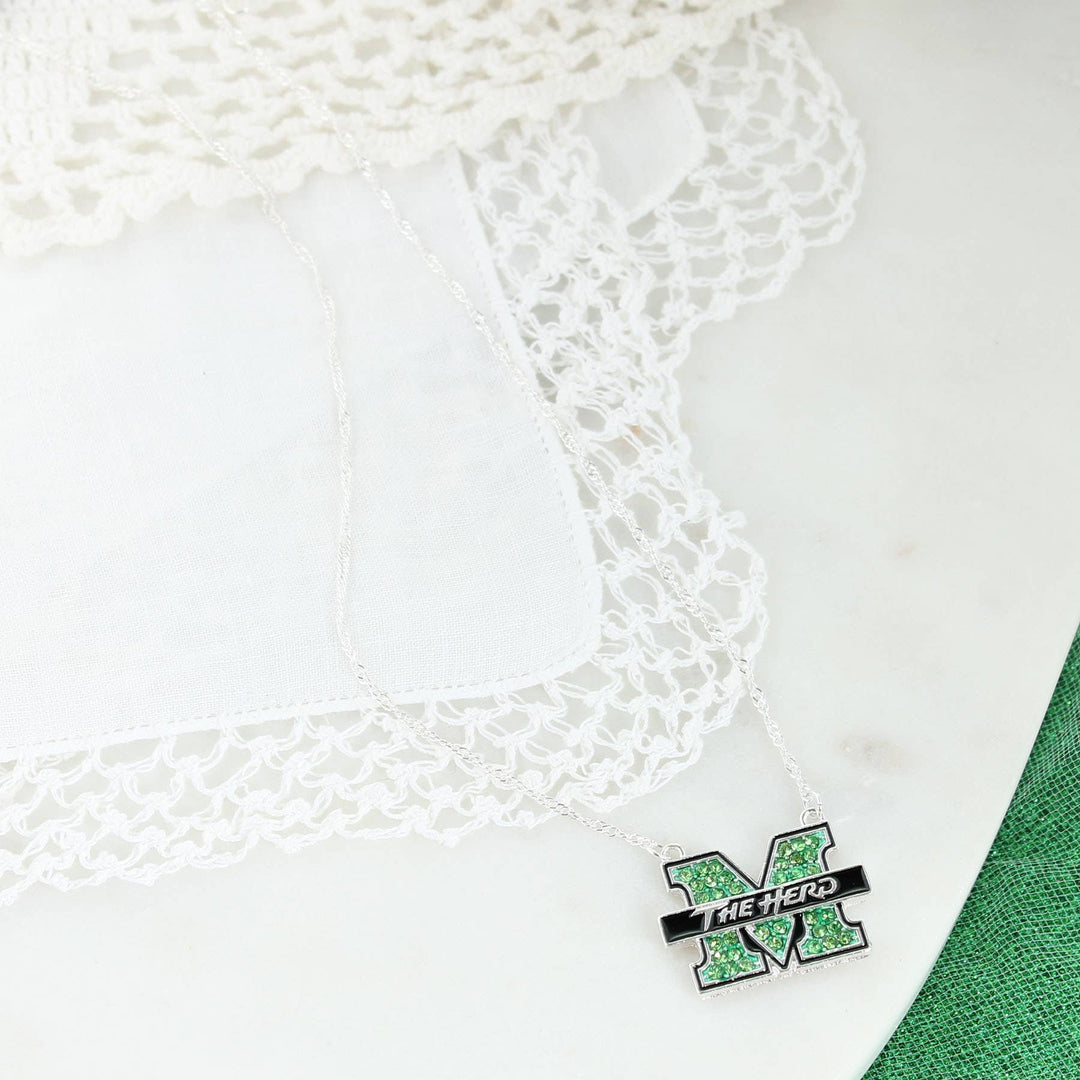 16” in length, this silver plated Marshall logo necklace features green rhinestones and black enamel with a claw clasp and a 3” extender. resting on white lacy background