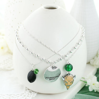 Set of three silver plated 16” necklaces (each with a claw clasp and 3” extender). Each necklace features a different chain style and charm, to include green and black beads, an enamel Marshall logo charm, and a silver stamped disc featuring the “Thundering Herd” slogan. On white vase